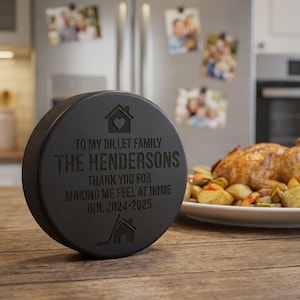 May include: A black hockey puck with engraved text. The text reads "TO MY BILLET FAMILY THE HENDERSONS THANK YOU FOR MAKING ME FEEL AT HOME OHL 2024-2025". The puck is on a wooden surface, with a meal in the background.