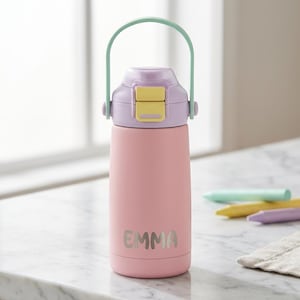 Könnte beinhalten: Eine rosa isolierte Wasserflasche mit hellgrünem Griff und lila Deckel mit gelbem Verschluss. Der Name "EMMA" ist in silbernen Buchstaben auf die Flasche gedruckt. Die Flasche ist für Kinder konzipiert.
