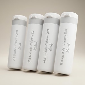 May include: Four white insulated water bottles with gray accents. Each bottle has a name and the text "With Gratitude - Volunteer 2024" printed on it. The names are Sarah, Michael, Emily, and David. The bottles are cylindrical with a flip-top lid.
