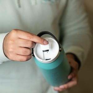 Close-up of teal hydro flask water bottle with white lid, being opened by hand. Perfect for sports, travel, and everyday hydration. Customizable options available.