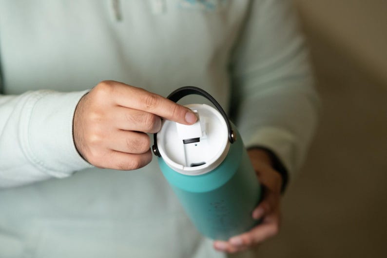 Close-up of teal hydro flask water bottle with white lid, being opened by hand. Perfect for sports, travel, and everyday hydration. Customizable options available.