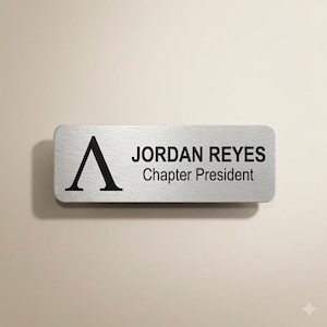 May include: A rectangular silver-toned name badge with a brushed metal finish. The badge features a large black letter A on the left side, followed by the text JORDAN REYES and the title Chapter President printed in a clean black sans-serif font.