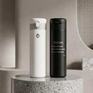 Op de afbeelding: Een witte geïsoleerde waterfles met een grijze dop en een waterdruppelpictogram, naast een zwarte geïsoleerde waterfles met de tekst: TODAY'S GOAL, CONTROL AIR TRAFFIC, WITHOUT YELLING ANYONE, TO GO F---K THEMSELVES, -TS.