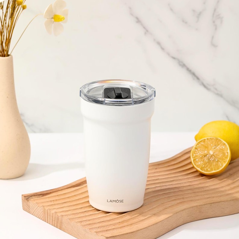 White stainless steel tumbler with clear lid, perfect for hot or cold beverages. Features Lamose branding. Includes sliced lemon on a wooden tray.