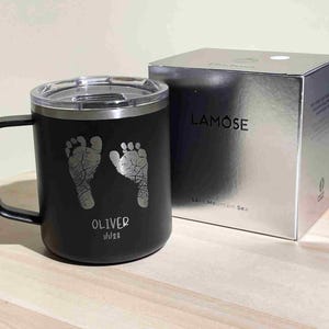 May include: Black travel mug with a silver rim and handle, featuring two silver baby footprints, the name "Oliver", and the date 1/1/23. A silver box with the word "LAMOSE" is next to the mug.