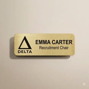 Peut inclure: Un badge nominatif rectangulaire de couleur dorée aux coins arrondis. Le côté gauche présente un logo triangulaire noir au-dessus du mot DELTA. Le côté droit affiche le nom EMMA CARTER en gras noir, avec Recruitment Chair imprimé en dessous.