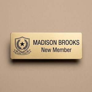 May include: A rectangular gold-toned name badge with a brushed metal finish. The badge features a black printed shield emblem with a star and laurel leaves, the text LAUREL SOCIETY, and the name MADISON BROOKS above the text New Member.