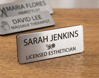 May include: A rectangular brushed silver name badge with black engraved text reading SARAH JENKINS, a small flower icon, and LICENSED ESTHETICIAN. Two additional blurred silver name badges are visible in the background with various names and titles.