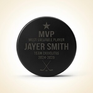 May include: A black hockey puck with engraved text. The text reads "MVP MOST VALUABLE PLAYER JAYER SMITH TEAM CRIIOLERS 2024-2025" with a star and crossed hockey sticks. A personalized sports award.