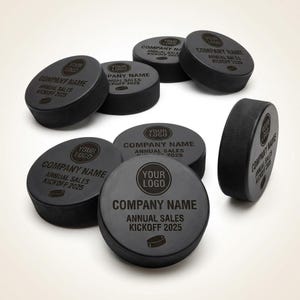 May include: Black hockey pucks featuring the text "YOUR LOGO", "COMPANY NAME", and "ANNUAL SALES KICKOFF 2025". The pucks are grouped together, with one positioned on its edge.