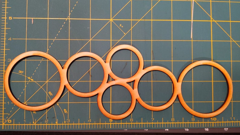 May include: Five wooden circles of varying sizes are arranged in a pattern on a green cutting mat. The circles are all the same color, a light orange-brown.