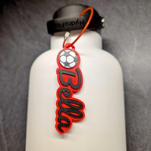 May include: Red and black personalized water bottle charm with the name "Bella" and a soccer ball graphic.