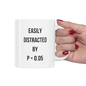 May include: White ceramic mug with the text "EASILY DISTRACTED BY P < 0.05" in black. The mug is held by a hand with red nail polish. The mug has a standard handle and is against a white background.