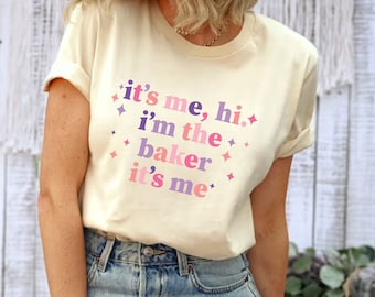 Peut inclure: Un t-shirt beige clair avec le texte "it's me, hi. i'm the baker it's me" en lettres roses, violettes et bleues avec des étoiles roses et violettes.