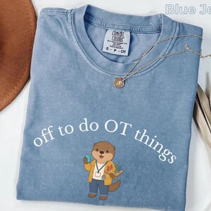 Könnte beinhalten: Ein kurzärmeliges T-Shirt in Jeansblau mit Rundhalsausschnitt. Das Shirt zeigt den weißen Schriftzug "off to do OT things" über einem gezeichneten Otter, der eine gelbe Strickjacke, eine blaue Hose und ein Schlüsselband trägt und ein buntes Steckspiel hält.