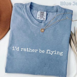 Peut inclure: Un t-shirt bleu jean avec l'inscription blanche "i'd rather be flying". Un collier doré avec un pendentif est posé sur le t-shirt. Le t-shirt est plié proprement sur une surface blanche.