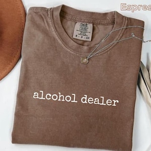 May include: A brown short-sleeved t-shirt with the text alcohol dealer printed in white on the front. The shirt features a crew neckline and a Comfort Colors brand label on the inside collar. The fabric has a washed, muted brown appearance.