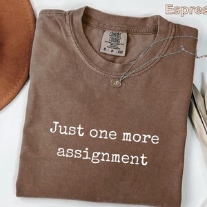 May include: An espresso-colored short-sleeve t-shirt with a crew neckline. The shirt features the white text "Just one more assignment" printed on the front. A silver-toned necklace with a small circular pendant rests on the collar area.