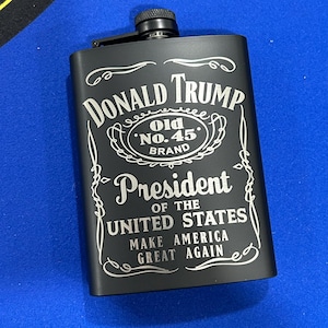 May include: Black stainless steel hip flask with a silver engraved design. The flask features the text "DONALD TRUMP Old 'No. 45' BRAND President OF THE UNITED STATES MAKE AMERICA GREAT AGAIN".