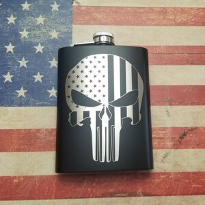 May include: Black stainless steel flask with a silver engraved design of a skull with an American flag pattern.