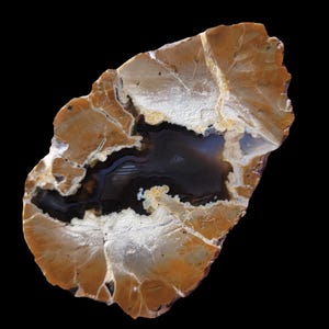 Peut inclure: Une grande géode d'agate de forme irrégulière avec un motif en bandes brun, blanc et noir. La géode a une grande cavité ouverte remplie d'une matière sombre et vitreuse.