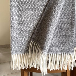 Puede incluir: Una manta tejida gris y blanca con un patrón geométrico y flecos color crema. La manta está sobre una silla de madera, mostrando su textura y diseño. Un elemento de decoración del hogar acogedor.