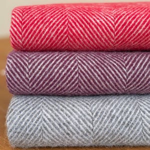 May include: Three rolls of fabric in red, purple, and grey, stacked on top of each other. Each roll features a herringbone design. The words "RED", "PURPLE", and "GREY" are printed on the left side. The fabric is likely woven.