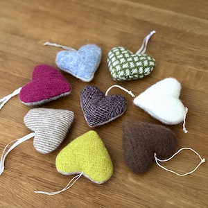 Könnte beinhalten: Eine Sammlung herzförmiger Ornamente in verschiedenen Farben und Texturen, darunter Blau, Grün, Lila und Braun. Jedes Herz hat eine weiße Schnur zum Aufhängen. Die Ornamente sind auf einer Holzoberfläche angeordnet.