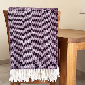 May include: A purple and white herringbone throw blanket with white fringe is draped over a wooden chair. The blanket is made of a soft, textured material. A white lamp sits on a wooden table in the background.