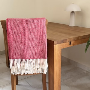 Op de afbeelding: Een rood-wit visgraatpatroon deken met witte franjes is over een houten stoel gedrapeerd. De deken is een levendig accent tegen de natuurlijke houttinten van de stoel en de tafel. Een witte lamp en potplanten zijn ook zichtbaar.