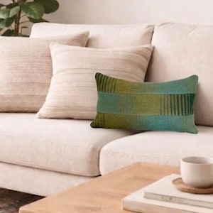 May include: A cream-colored sofa with three decorative pillows and a rectangular green and yellow patterned pillow. A wooden side table with a lamp and a coffee table with a cup and saucer are in front of the sofa.