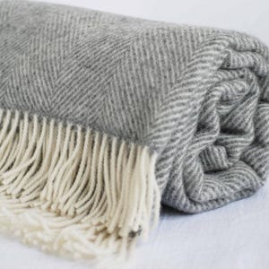 Op de afbeelding: Opgerolde grijze en witte visgraat plaid met crèmekleurige franjes. De deken, gemaakt van een zacht, getextureerd materiaal, toont zijn patroon en franjes. De neutrale kleuren passen bij verschillende interieurstijlen. De deken is opgerold.