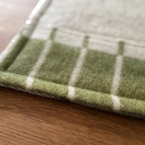 K&ouml;nnte beinhalten: Nahaufnahme eines gr&uuml;n-wei&szlig; karierten Textils. Das Textil hat ein weiches, strukturiertes Aussehen mit einer gr&uuml;nen Basis und wei&szlig;en vertikalen Streifen. Die Kanten sind sauber mit einem wei&szlig;en Rand versehen. Der Artikel ist wahrscheinlich ein K&uuml;chen- oder Wohnaccessoire.