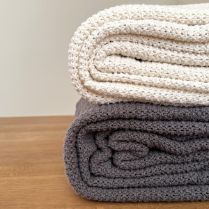 Peut inclure: Deux couvertures pliées empilées l'une sur l'autre. La couverture du dessus est de couleur crème avec un motif en maille gaufrée texturée. La couverture du dessous est gris foncé et présente la même texture en maille gaufrée uniforme.