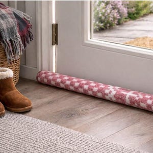 Op de afbeelding: Een rood-witte geblokte tochtstrip ligt op een houten vloer bij een deur. Bruine suède laarzen met witte bontafwerking staan op een grijze vloerkleed. Een rieten mand bevat een geruite deken.