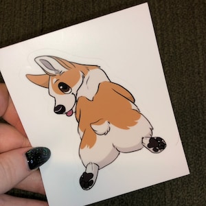 May include: A white sticker featuring a cartoon corgi dog, viewed from behind, looking over its shoulder. The corgi has brown and white fur, with black paws and a pink tongue sticking out. The sticker is held against a green background.