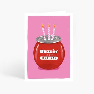 May include: A birthday card with a red can design, featuring the text "Buzzin' it's your BIRTHDAY". Three striped candles with lit flames sit atop the can against a pink background.