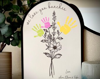 May include: A white wooden sign with a black frame in the shape of an arch. The sign has a floral design with three handprints in pink, yellow, and green. The text "we love you bunches" is written at the top of the sign. The text "Love, Ava, Olivia & Zoey" is written at the bottom of the sign.