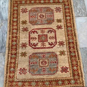 Könnte beinhalten: Rechteckiger beigefarbener Teppich mit einem detaillierten geometrischen Muster in Rot, Braun und Blau. Der Teppich hat ein zentrales Design mit sich wiederholenden floralen und geometrischen Motiven und einen Fransenrand. Der Teppich ist für ein Wohn- oder Schlafzimmer geeignet.