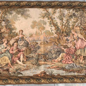 Puede incluir: Un tapiz que representa una escena pastoral con figuras en un paisaje boscoso. El tapiz presenta un grupo de personas pescando en un arroyo, con árboles y follaje que los rodean. El tapiz está enmarcado en un marco dorado con un borde decorativo.