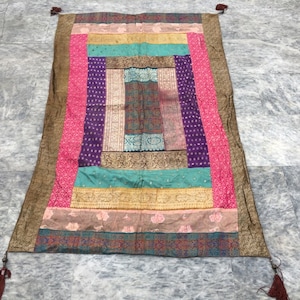 Könnte beinhalten: Eine bunte Patchwork-Decke mit braunem Rand und Quasten. Die Decke besteht aus verschiedenen Stoffen und Mustern, darunter Rosa, Lila, Türkis und Gold.