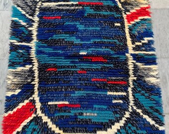 Puede incluir: Una alfombra abstracta azul, roja y blanca con una textura peluda. La alfombra tiene una gran forma ovalada en el centro con un diseño geométrico.