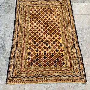 Puede incluir: Una alfombra rectangular, tejida a mano, con un campo central amarillo y patrones repetidos en forma de hojas marrones. La alfombra tiene un borde detallado en tonos marrones y rojos, con una franja marrón oscuro.