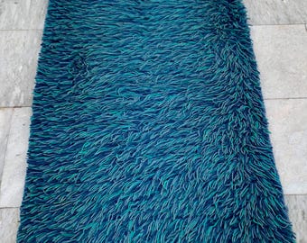 Puede incluir: Una alfombra rectangular y peluda en tonos turquesa y azul. La alfombra tiene un pelo denso y texturizado, creando una apariencia suave y acogedora. El color transita de un azul profundo en los bordes a un turquesa más claro en el centro.