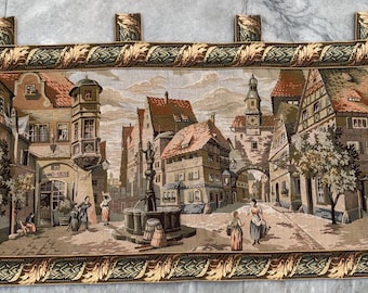 Könnte beinhalten: Ein Wandteppich, der einen europäischen Stadtplatz mit Kopfsteinpflasterstraßen, Gebäuden mit roten Ziegeldächern und einem zentralen Brunnen zeigt. Der Wandteppich hat einen dekorativen Rand mit Blattmustern und Aufhängeösen.
