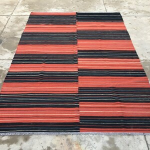 Pu&ograve; includere: Un tappeto a strisce rosse e nere con un bordo a frange. Il tappeto &egrave; fatto di tessuto tessuto e ha un design tradizionale.