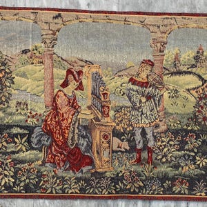Puede incluir: Tapiz que representa una escena renacentista con figuras tocando instrumentos en un jardín. El tapiz presenta un borde rojo y un paisaje detallado con elementos florales y arquitectónicos.