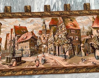 May include: A tapestry depicting a European village scene with colorful buildings, cobblestone streets, and people walking. The tapestry has a brown frame with tassels.