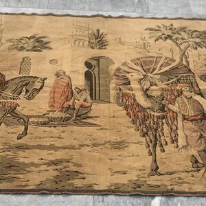Op de afbeelding: Een vintage wandtapijt dat een scène voorstelt van mensen die op kamelen en paarden rijden in een woestijnlandschap. Het wandtapijt is geweven met ingewikkelde details en heeft een verscheidenheid aan kleuren, waaronder beige, bruin en wit.