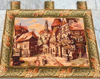 Könnte beinhalten: Ein Wandteppich mit einem grünen und goldenen Rahmen, der eine europäische Dorfszene mit Gebäuden, einem Brunnen und zwei Figuren darstellt. Der Wandteppich ist aus gewebtem Stoff gefertigt und hat ein detailliertes Design.